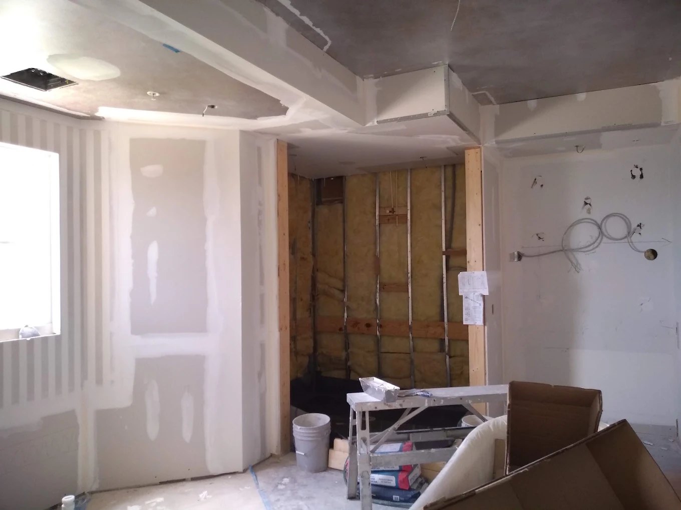 Interior renovation in progress with drywall, exposed insulation, plastic sheeting, and construction materials